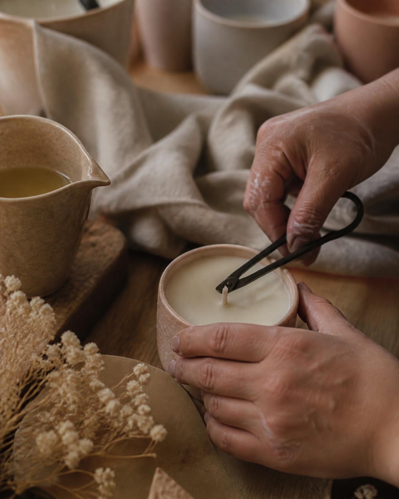 Mãos do artesão ajustando o pavio de uma vela artesanal em recipiente de cerâmica sobre bancada de madeira